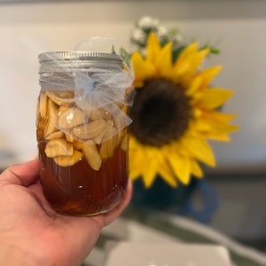 May include: A glass jar filled with honey and sliced garlic, tied with a white ribbon and a mesh bag. The jar is being held in a hand, with a sunflower in the background.