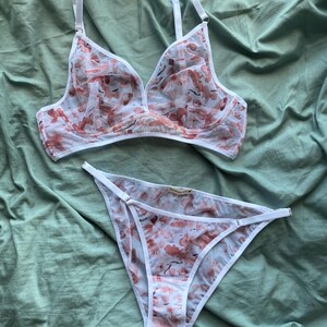 Könnte beinhalten: Ein Dessous-Set, bestehend aus einem BH und einem passenden Slip. Das Set ist weiß mit einem zarten Blumenmuster in Rosa- und Pfirsichtönen. Der BH hat verstellbare Träger und ein transparentes Design.