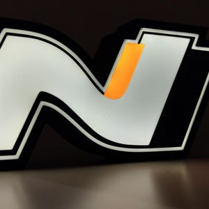 May include: Illuminated black and white 'N' logo with an orange accent. The logo is lit from within, creating a bright glow against a dark background. The design features a bold, modern aesthetic, suitable for automotive or branding applications.
