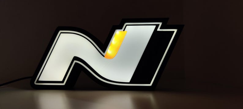May include: Illuminated sign featuring the letter "N" in a stylized design. The sign has a black border with a white and silver interior, and a yellow accent. The sign is lit, creating a glowing effect.