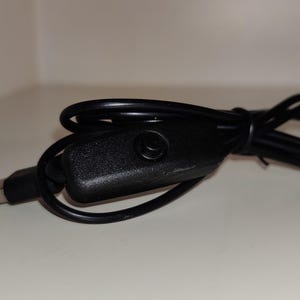 May include: A black USB cable with a rectangular, textured switch and a USB-A connector. The cable is coiled and lies on a white surface. The switch has a small circular button.