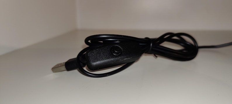 May include: A black USB cable with a rectangular, textured switch and a USB-A connector. The cable is coiled and lies on a white surface. The switch has a small circular button.