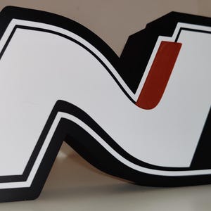 May include: A large, stylized letter "N" in black, white, and red. The letter has a thick black border with a white interior and a red vertical stripe. The design is likely a logo or emblem, possibly for a brand or organization.