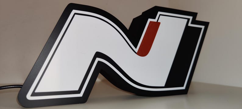 May include: A large, stylized letter "N" in black, white, and red. The letter has a thick black border with a white interior and a red vertical stripe. The design is likely a logo or emblem, possibly for a brand or organization.