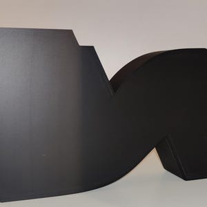 May include: A large, matte black letter "M" sculpture with a modern design. The sculpture has a geometric shape with a flat, angled side and a curved section. A black power cord extends from the sculpture.
