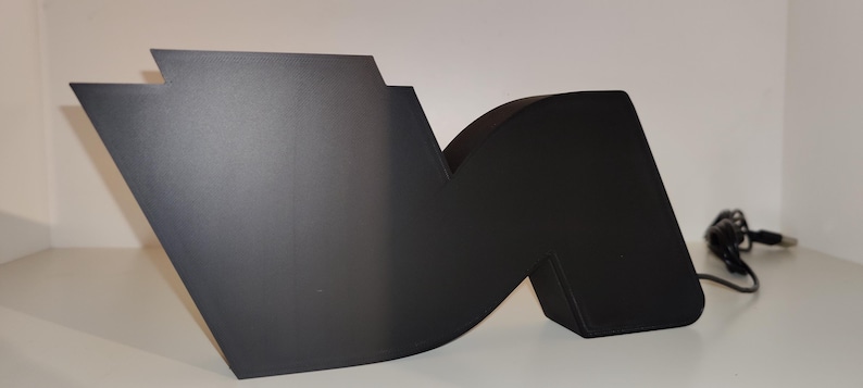 May include: A large, matte black letter "M" sculpture with a modern design. The sculpture has a geometric shape with a flat, angled side and a curved section. A black power cord extends from the sculpture.