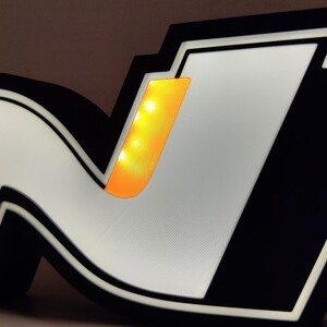 May include: Illuminated sign featuring the letter "N" in a stylized design, with a black border and a white interior. The "N" has a yellow-orange accent. The sign is lit, creating a glowing effect.