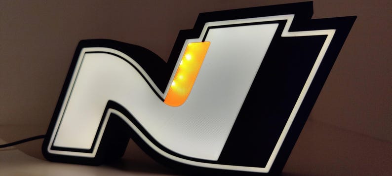 May include: Illuminated sign featuring the letter "N" in a stylized design, with a black border and a white interior. The "N" has a yellow-orange accent. The sign is lit, creating a glowing effect.