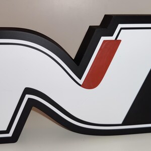 May include: A large, illuminated sign in the shape of the letter "N". The sign features a white interior with a black outline and a red accent. The background is white.