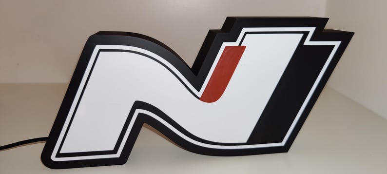 May include: A large, illuminated sign in the shape of the letter "N". The sign features a white interior with a black outline and a red accent. The background is white.