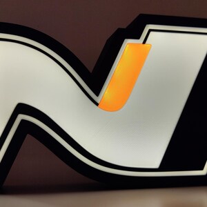 May include: Illuminated sign featuring a stylized white "N" logo with a black outline and accents. The design includes an orange vertical bar within the "N". The sign is lit, creating a bright, eye-catching display. The overall aesthetic is modern and graphic.