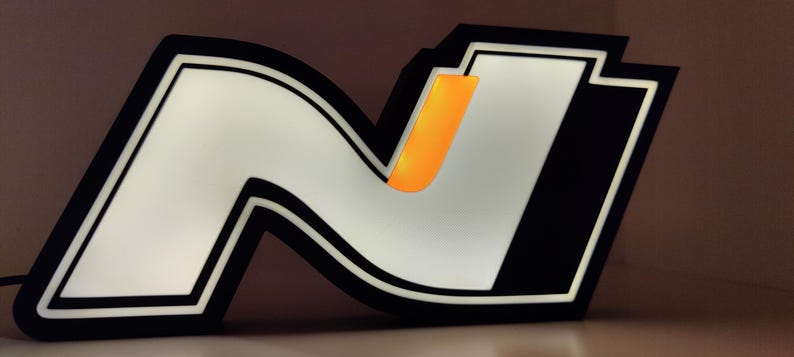 May include: Illuminated sign featuring a stylized white "N" logo with a black outline and accents. The design includes an orange vertical bar within the "N". The sign is lit, creating a bright, eye-catching display. The overall aesthetic is modern and graphic.