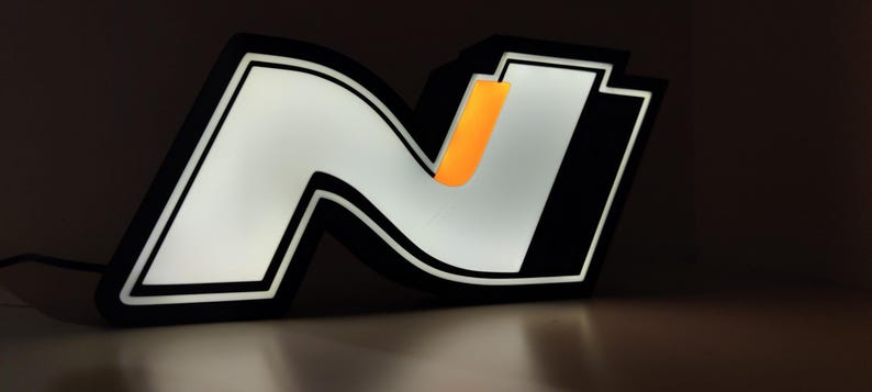 May include: Illuminated black and white 'N' logo with an orange accent. The logo is lit from within, creating a bright glow against a dark background. The design features a bold, modern aesthetic, suitable for automotive or branding applications.