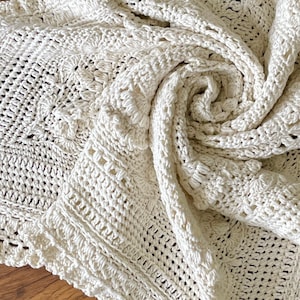 Może przedstawiać: Koc wykonany na szydełku w kremowym kolorze z delikatnym, skomplikowanym wzorem. Koc jest złożony i drapowany na drewnianej powierzchni.
