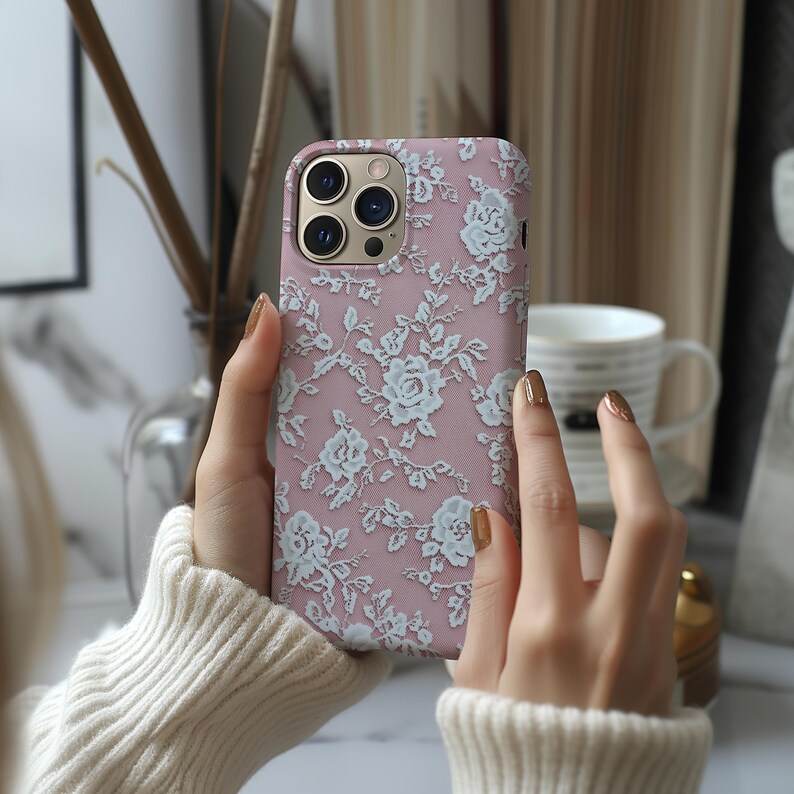May include: A pink phone case with a white lace pattern. The case has a camera lens cutout and a cutout for the phone's buttons.
