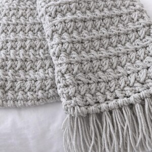 May include: A light gray, hand-crocheted scarf with a textured stitch pattern and fringe detail. The scarf is made of soft yarn and is perfect for adding warmth and style. The scarf is approximately 70 inches long.