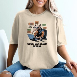 May include: Beige t-shirt with a graphic of an anteater wearing glasses and coding on a laptop. The text "CODE, EAT, SLEEP, REPEAT" is printed below the anteater. The shirt is a relaxed fit.