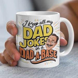 May include: White ceramic coffee mug with a cartoon illustration of a man with a mustache and glasses. The mug has the text "I keep all my DAD JOKES in a DAD-A-BASE" in yellow and orange lettering.