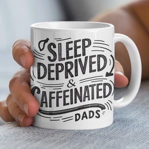 Op de afbeelding: Een witte keramische koffiemok met zwarte tekst die "Sleep Deprived & Caffeinated Dads" luidt.