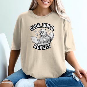 Pode incluir: Camiseta bege com um gráfico em preto e branco de um castor de óculos trabalhando em um laptop. O texto "CODE, BUILD, REPEAT" está acima e abaixo da ilustração do castor. A camiseta tem um caimento folgado.