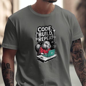 Könnte beinhalten: Graues T-Shirt mit einem Cartoon-Biber, der ein rotes Hemd trägt und an einem grünen Laptop arbeitet. Der Text "CODE, BUILD, REPEAT" steht über dem Biber. Das T-Shirt ist ein lässiger Stil.