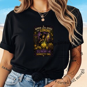 Può includere: Una t-shirt nera con una stampa grafica di una strega che mescola un calderone giallo brillante. Il testo recita: WHEN LIFE GIVES YOU LEMONS SUMMON SOMETHING. Il design include teschi, un gatto nero e un cartello con scritto CURSED LEMONADE.
