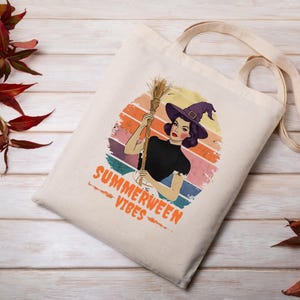 Puede incluir: Bolso tote de color crema con una ilustraci&oacute;n retro de una bruja sosteniendo una escoba. El dise&ntilde;o incluye un fondo de puesta de sol colorido y las palabras "SUMMERWEEN VIBES" en naranja.