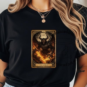 May include: Black t-shirt featuring a tarot card design. The card depicts a demonic figure with horns and wings, surrounded by flames. The card is framed in gold with the text "THE DEMON".