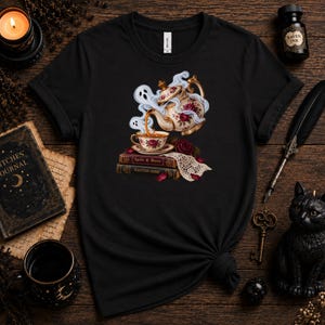 May include: A black short-sleeve t-shirt featuring a graphic of a floral teapot pouring tea into a matching cup, resting on two books titled Spells & Brews and Haunted Home, surrounded by swirling ghost-shaped steam and a piece of white lace.
