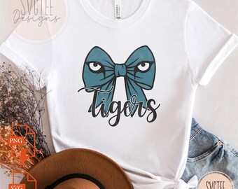 May include: White t-shirt featuring a teal bow design with eyes and the word "Tigers" in black script. The shirt is made of a soft material and has short sleeves. The design is centered on the front of the shirt.