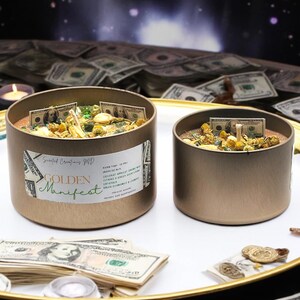 May include: Two gold candle jars with a golden manifest label on one jar. The jars are filled with dried herbs, flowers, and dollar bills. The jars are sitting on a white surface surrounded by stacks of dollar bills.