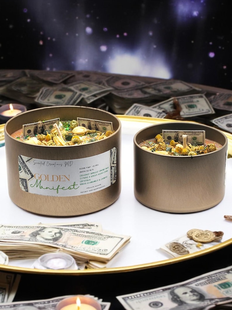 May include: Two gold candle jars with a golden manifest label on one jar. The jars are filled with dried herbs, flowers, and dollar bills. The jars are sitting on a white surface surrounded by stacks of dollar bills.