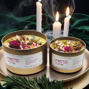 May include: Two gold candle jars with lids, labeled "Mystic Vision", filled with a green wax blend and topped with dried flowers and crystals. The candles are on a gold tray with a sprig of rosemary.