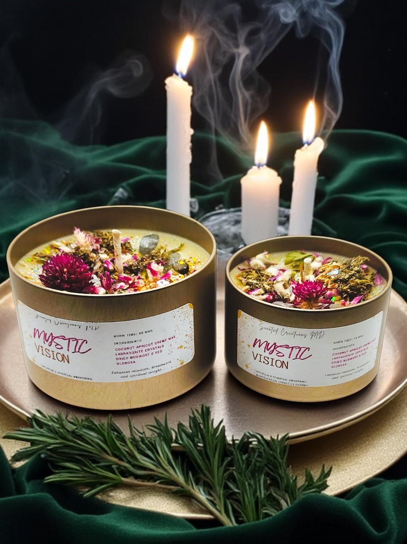 May include: Two gold candle jars with lids, labeled "Mystic Vision", filled with a green wax blend and topped with dried flowers and crystals. The candles are on a gold tray with a sprig of rosemary.