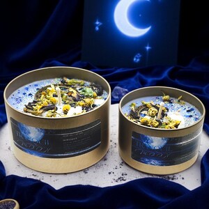 May include: Two gold candle jars with blue and white wax and dried flowers. The jars have a label that says "Tranquil Moon" and are on a blue velvet surface.