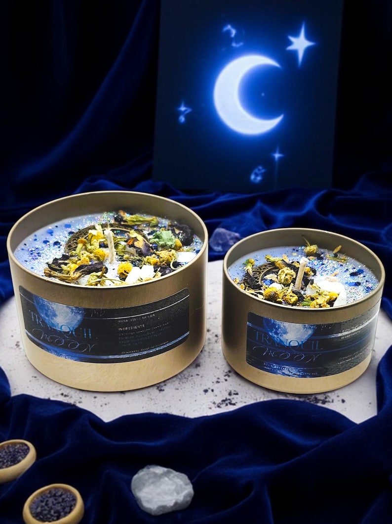 May include: Two gold candle jars with blue and white wax and dried flowers. The jars have a label that says "Tranquil Moon" and are on a blue velvet surface.