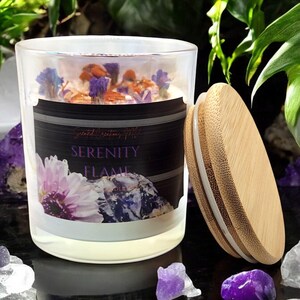May include: A glass candle jar with a white frosted finish and a black label that reads "Serenity Flame Candle Co.". The candle is topped with dried flowers and has a bamboo lid.