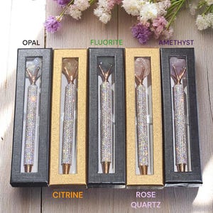 May include: Five black boxes with gold trim, each containing a crystal-topped pen. The boxes are labeled with the names of different crystals: Opal, Fluorite, Amethyst, Citrine, and Rose Quartz.