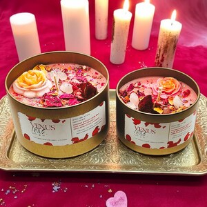 May include: Two gold candle jars with pink wax and dried flowers and crystals. The jars are labeled "Venus Love" and have a list of ingredients. The jars are on a gold tray with five white candles in the background.