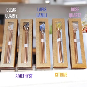 Puede incluir: Cinco cajas doradas con ventanas transparentes que muestran bolígrafos de cristal. Las cajas están etiquetadas con los nombres de los cristales: Cuarzo claro, Amatista, Lapislázuli, Citrino y Cuarzo rosa.
