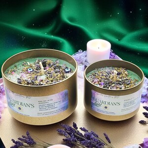 May include: Two gold candle jars with green wax and various crystals and herbs. The labels read "Guardian's Light" and list ingredients including coconut apricot creme wax, dried sage, and amethyst crystals. The candles are on a surface with lavender flowers and a white candle.