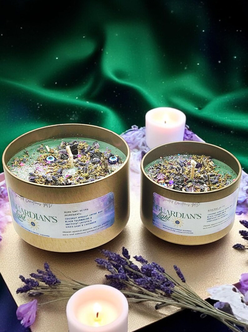 May include: Two gold candle jars with green wax and various crystals and herbs. The labels read "Guardian's Light" and list ingredients including coconut apricot creme wax, dried sage, and amethyst crystals. The candles are on a surface with lavender flowers and a white candle.