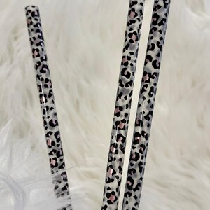 May include: Three cylindrical straws featuring a repeating leopard print pattern. The design consists of black and soft pink spots set against a translucent, frosted white background, creating a distinct animal-inspired aesthetic.