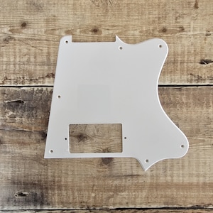 Puede incluir: Un golpeador de guitarra blanco con un recorte rectangular y varios agujeros para tornillos. El golpeador tiene una forma única con un borde curvo, sobre un fondo de madera.