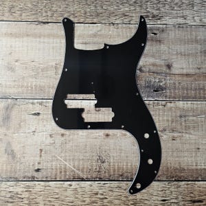 May include: Black plastic pickguard for a bass guitar. The pickguard has cutouts for the controls and pickups.