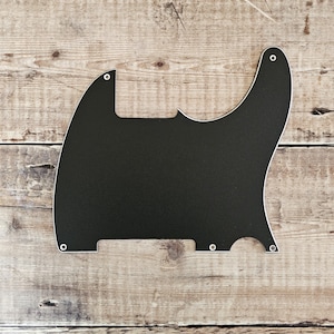 Może przedstawiać: Czarny pickguard do gitary z białym obramowaniem i otworami montażowymi. Pickguard ma unikalny kształt, zaprojektowany tak, aby pasował do konkretnego modelu gitary. Pickguard jest wykonany z solidnego, nieprzezroczystego materiału.