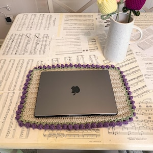 May include: A laptop rests on a crocheted placemat with a purple border. A white pitcher holds three crocheted tulips in yellow, pink, and purple. The table is covered with sheet music.