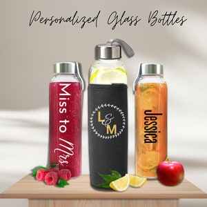 Puede incluir: Tres botellas de agua de vidrio con texto personalizado y una funda de neopreno negra. Las botellas están llenas de líquidos de diferentes colores. El texto en las botellas dice "Miss to Mrs", "L&M" y "Jessica".