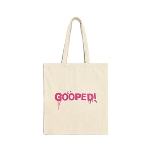 Puede incluir: Una bolsa de tela beige con un gráfico rosa que dice "GOOPED!" con efectos de pintura que gotean.