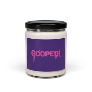 Puede incluir: Un tarro de vela blanco con tapa negra y una etiqueta morada que dice "GOOPED!" en letras rosas que gotean.